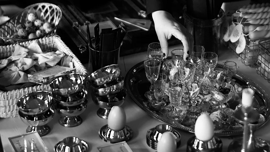Svart och vit bild. En hand sträcker sig över ett festligt dukat bord med små snaps glas på en silvrig bricka, en hög med snurrade kronljus, äggkoppar i silver med fake ägg placerade, en flätad fyrkantig korg med rosetter  