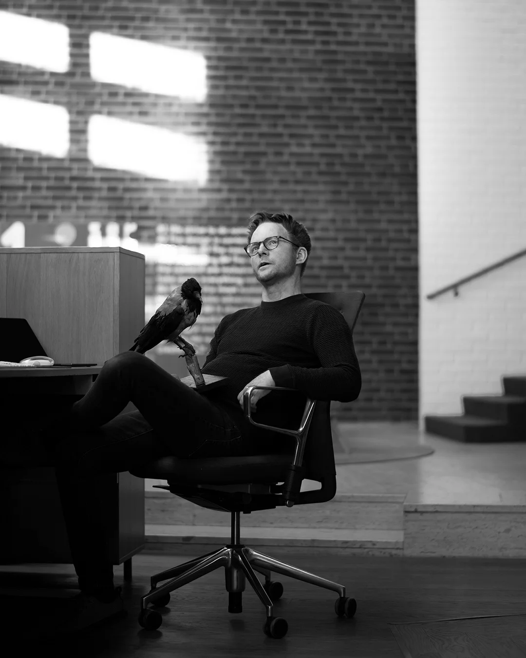 Svart och vit bild, man med glasögon sitter i en kontorsstol framför en tegelvägg. Huvudet kollar snett uppåt, funderande samtidigt som han håller i en uppstoppad kråka 