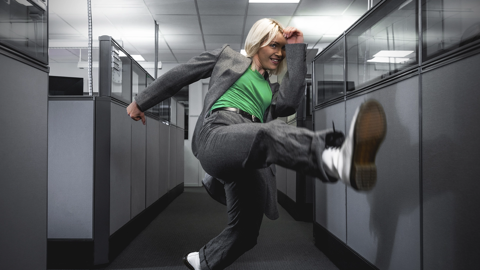  Woman in green t-shirt with suit jumping and kicking in the air, in an office. NyStudio