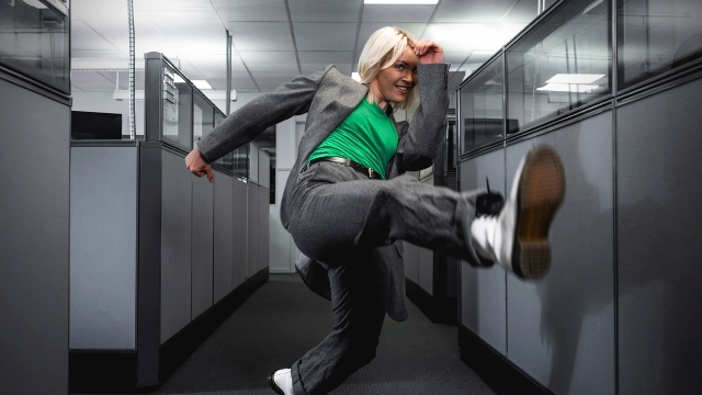 Woman in green t-shirt with suit jumping and kicking in the air, in an office. NyStudio