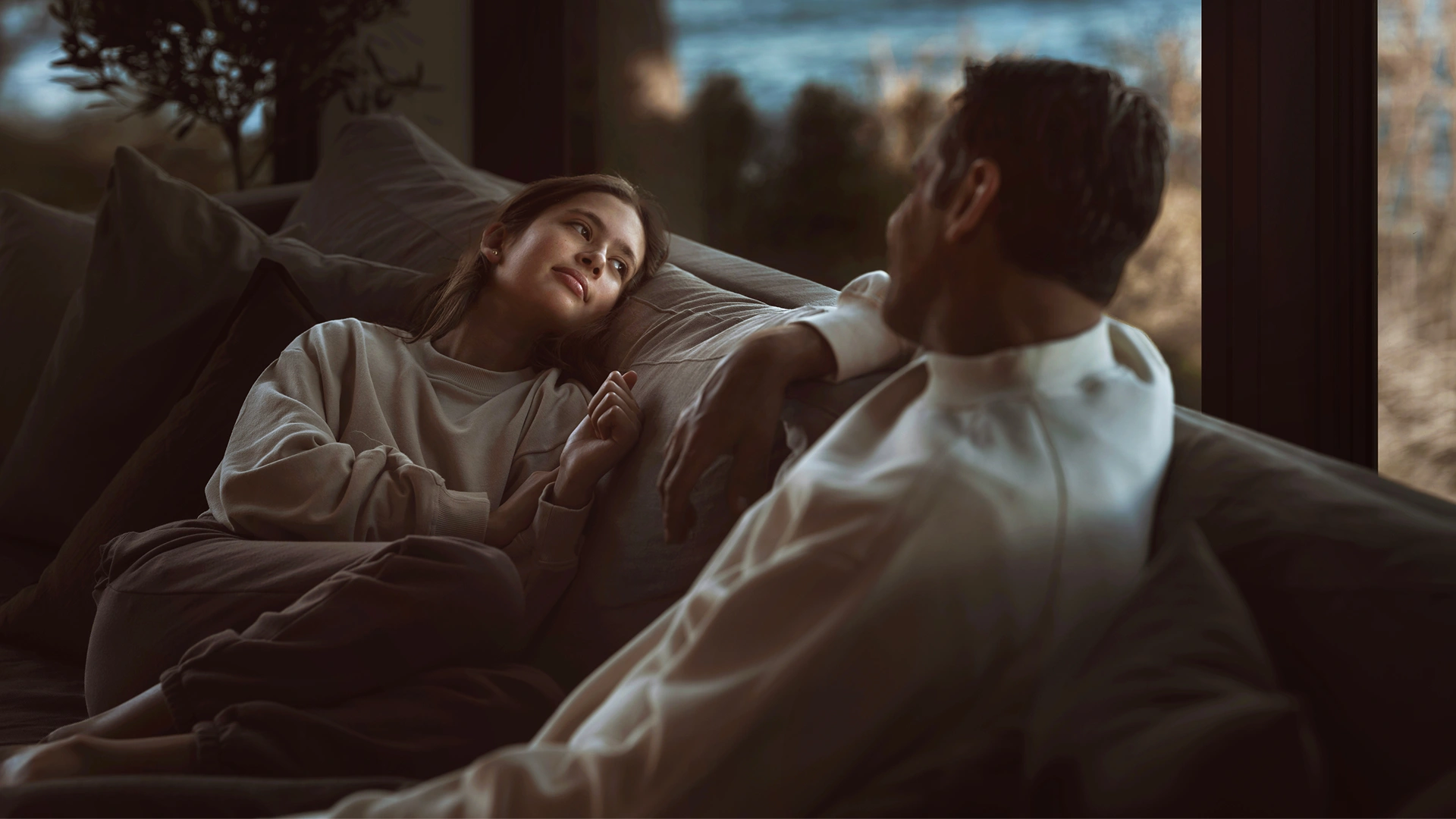 En man sitter på en soffa vänd mot en kvinna som ligger och tittar genom fönstret