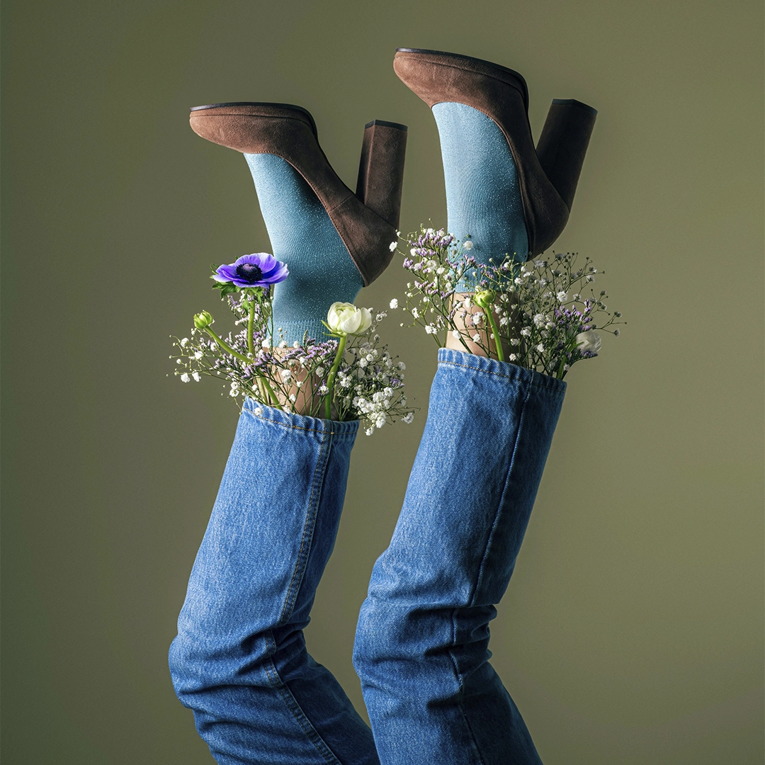 Brand image of a pair of legs with jeans, blue socks and brown heels and flowers around the ankles positioned in the air - NyStudio
